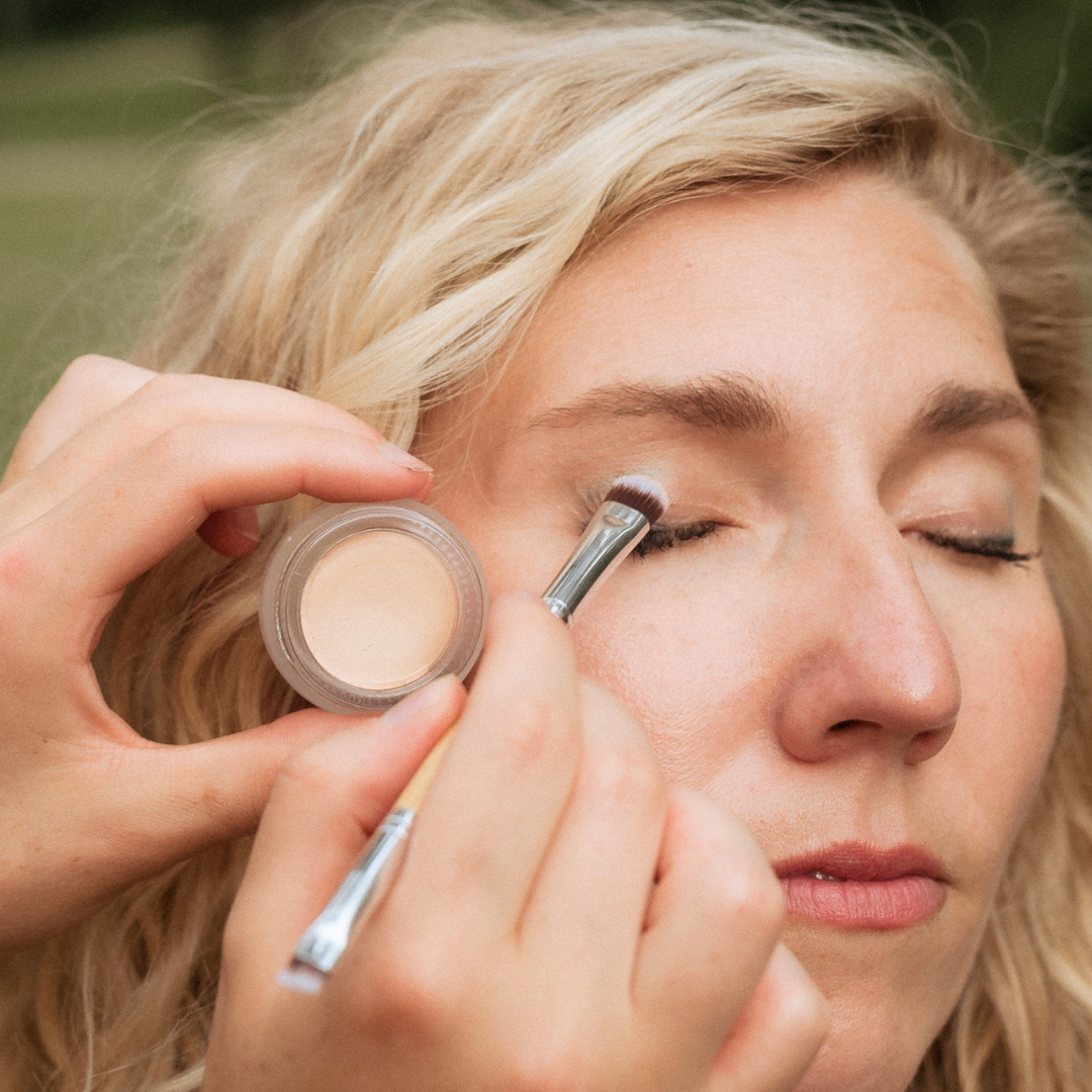 A close-up lifestyle shot of a woman applying concealer as a primer with a small makeup brush while holding an open pot of cream eyeshadow outdoors.