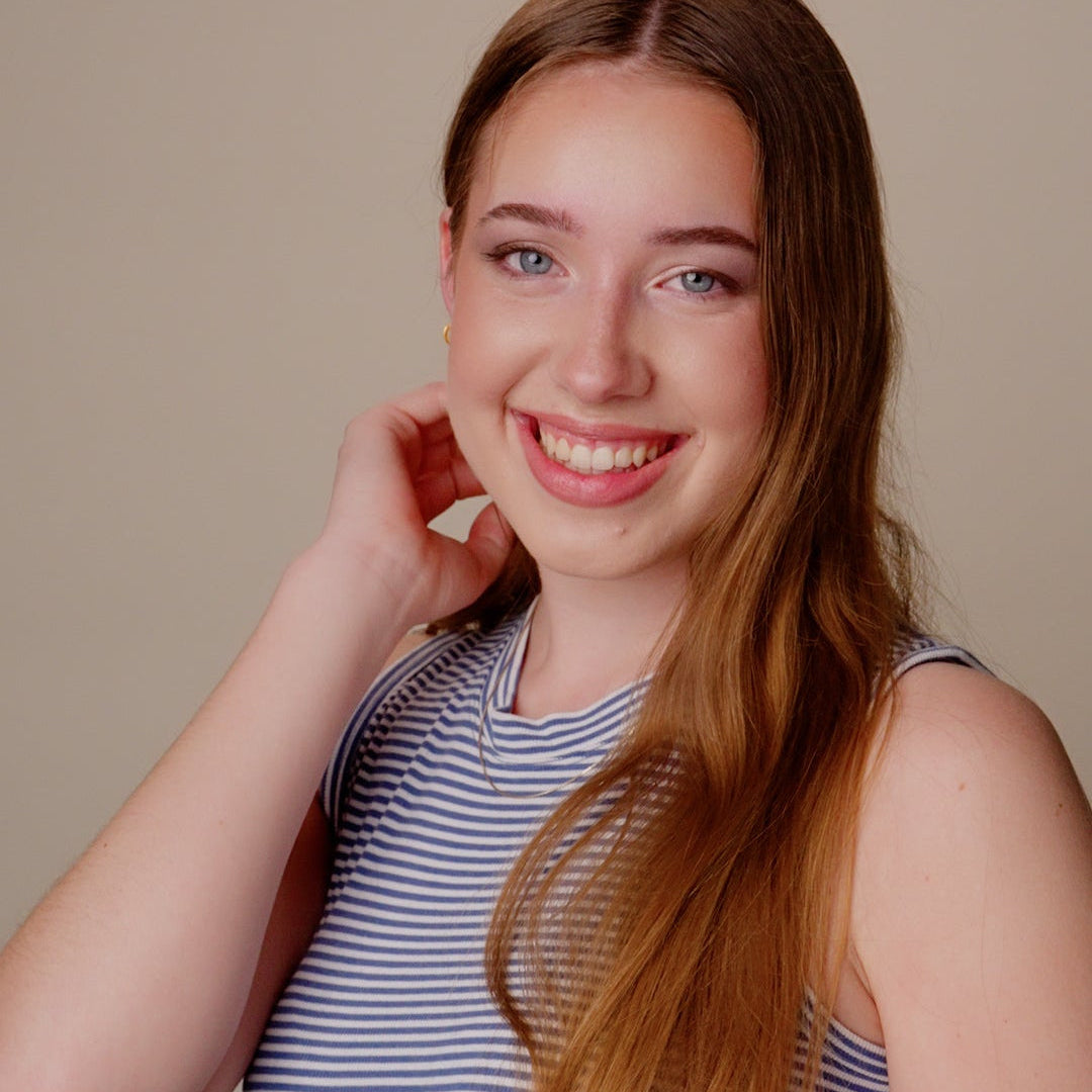 A smiling young woman with long light brown hair wearing a blue and white striped sleeveless top, posing against a solid beige background.