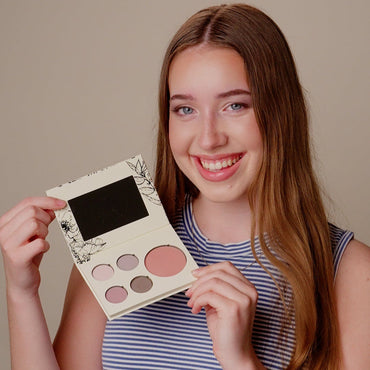 A smiling young woman with long light brown hair holds up a makeup palette with six pans of eyeshadow and blush. She is wearing a blue and white striped top against a solid beige background.
