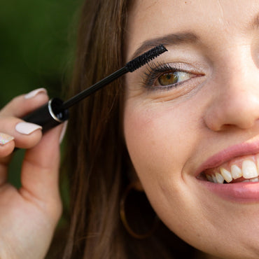 A close-up lifestyle shot of a woman applying black eyebrow mascara with a black wand. She has long brown hair, light skin, and is wearing gold hoop earrings, set against a soft, blurred green background.