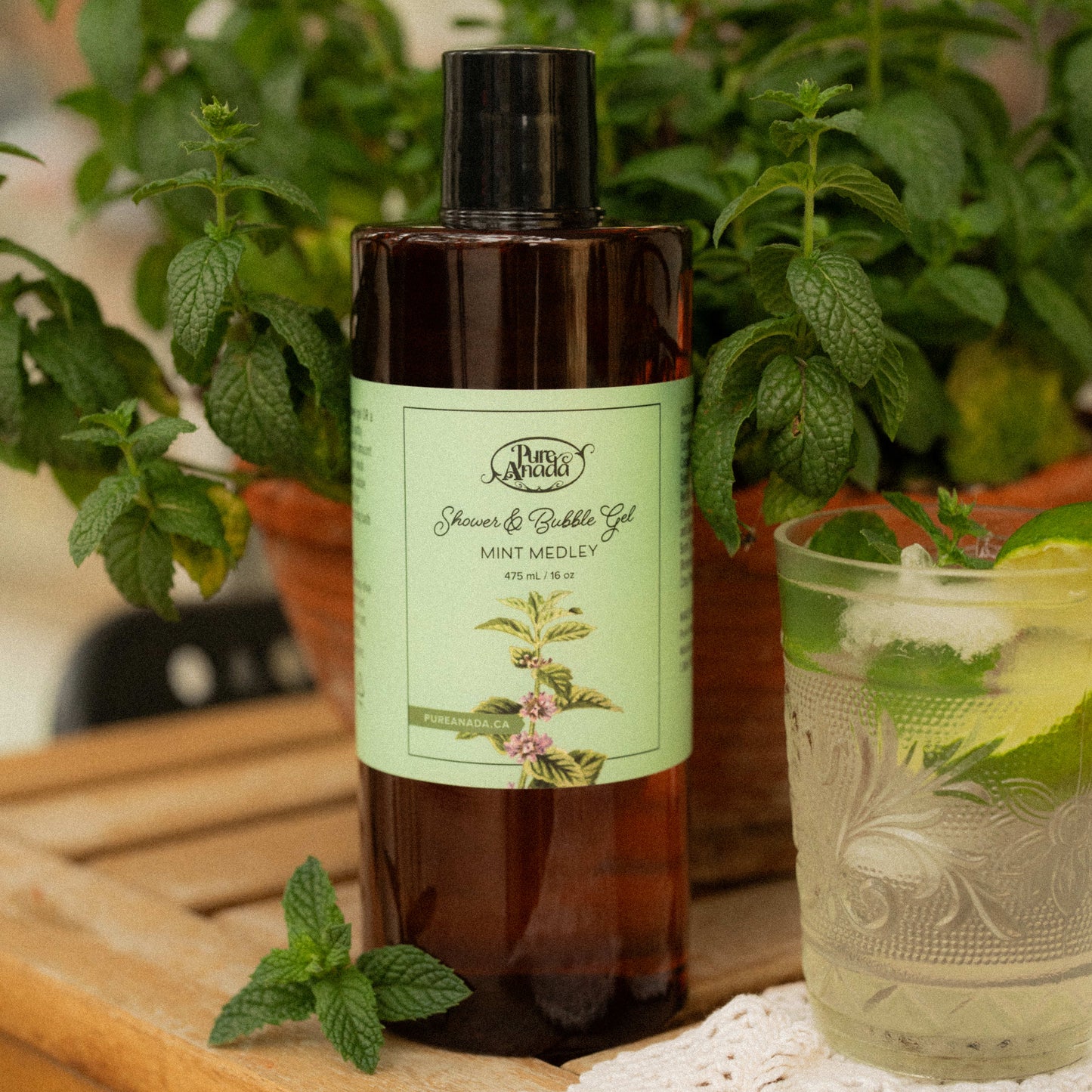 Bottle of 'Pure Anada' shower and bubble gel with a label, surrounded by mint leaves and a glass of lemonade.