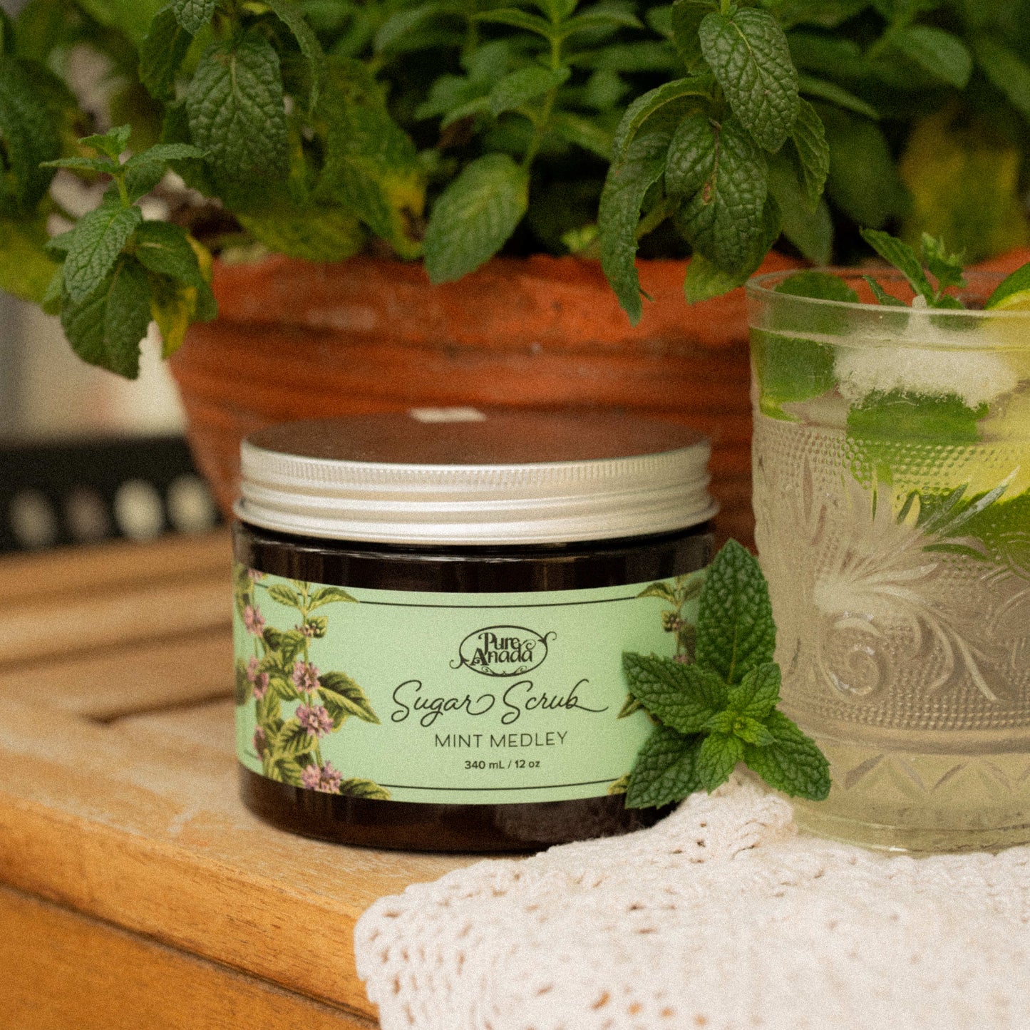 Jar of mint sugar scrub on a wooden surface with a potted mint plant and glass of water in the background.