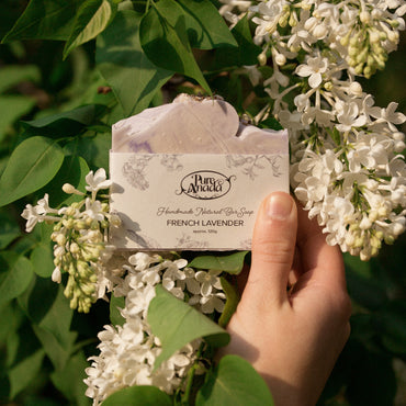 A hand holding a bar of soap with 'Pur Anada' brand name visible, against a background of white lavender flowers.