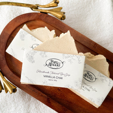 Handmade soap bar wrapped in paper, placed on a wooden dish with a white cloth background, labeled 'Vanilla Chai'.