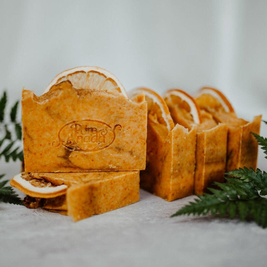 A photo of several bars of soap with a solid orange color and a visible exfoliating addition, displayed on a white surface with green leaf accents around it.