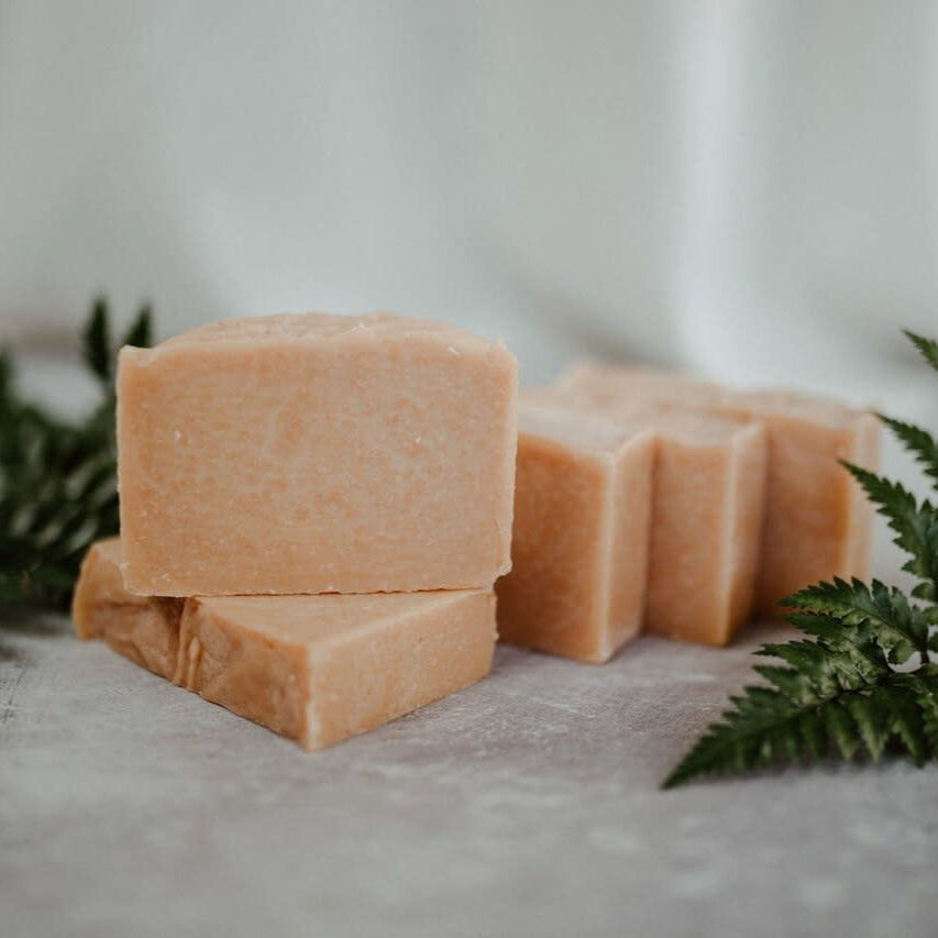 A bar of soap with a pink clay component, displayed on a grey surface with green leaves to the side.