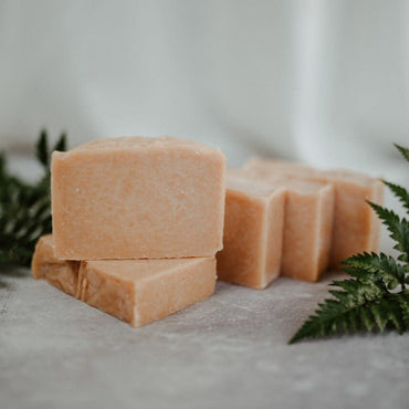 A bar of soap with a pink clay component, displayed on a grey surface with green leaves to the side.