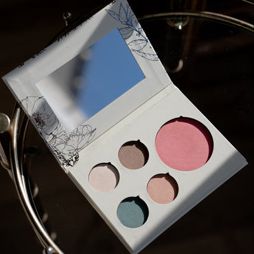 An open makeup palette with a mirror on the inside lid and five round pans in pink, mauve, peach, blue, and taupe, photographed on a dark reflective surface.