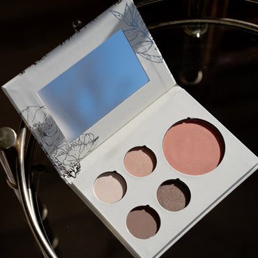 An open makeup palette with a large mirror on the inside lid and five round cosmetic pans in neutral and pink shades, placed on a dark reflective surface.