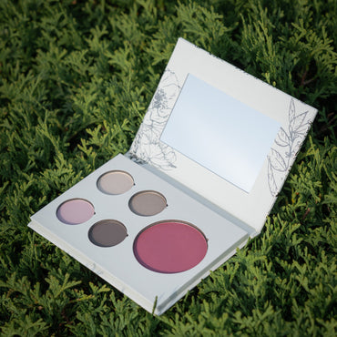 An open makeup palette with four circular and one oval powder pans in neutral and pink shades, resting on green foliage. The compact has a white cover with delicate floral line art and a mirror in the lid.