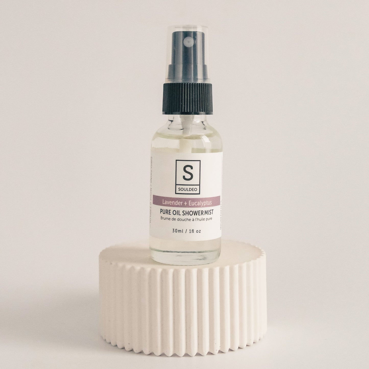 A clear spray bottle labeled SoulDeo with a white cap, containing a shower mist, placed on a white textured surface.