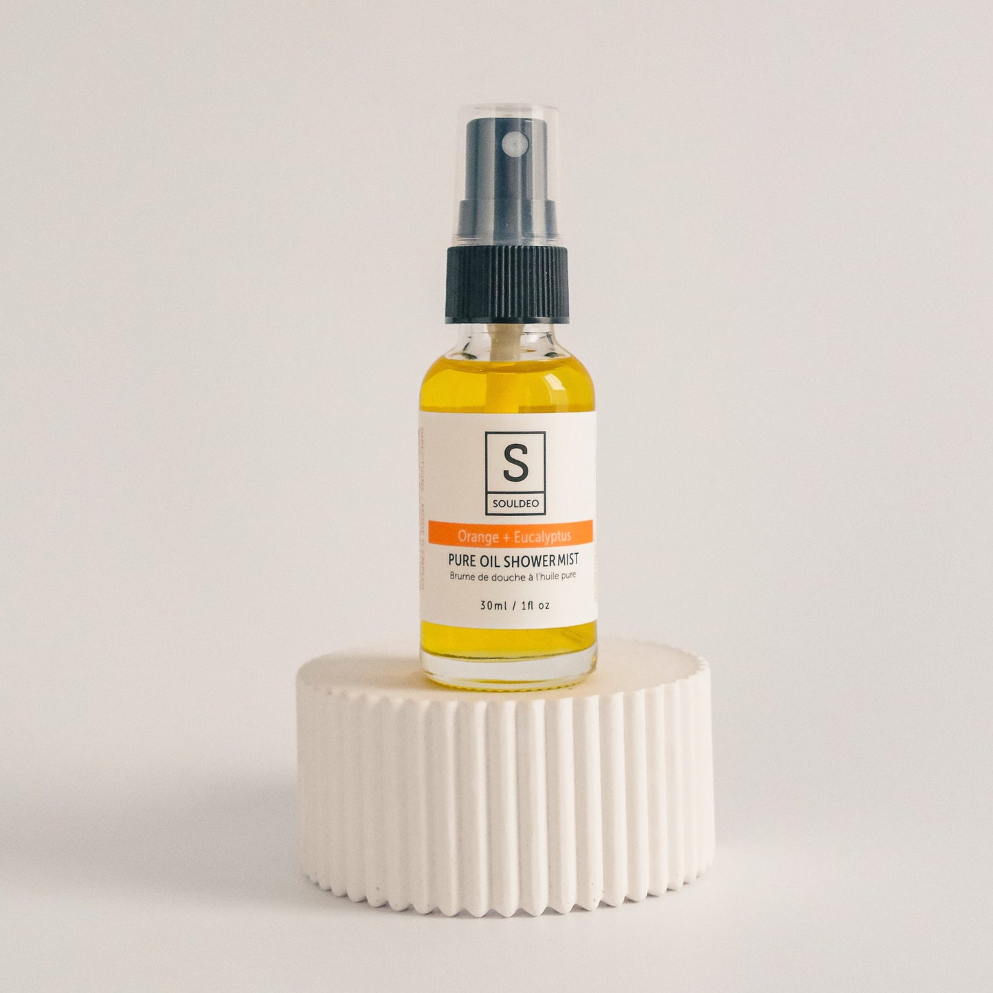 A small white spray bottle labeled SoulDeo Shower Mist with orange and eucalyptus, positioned on a textured white surface against a neutral background.