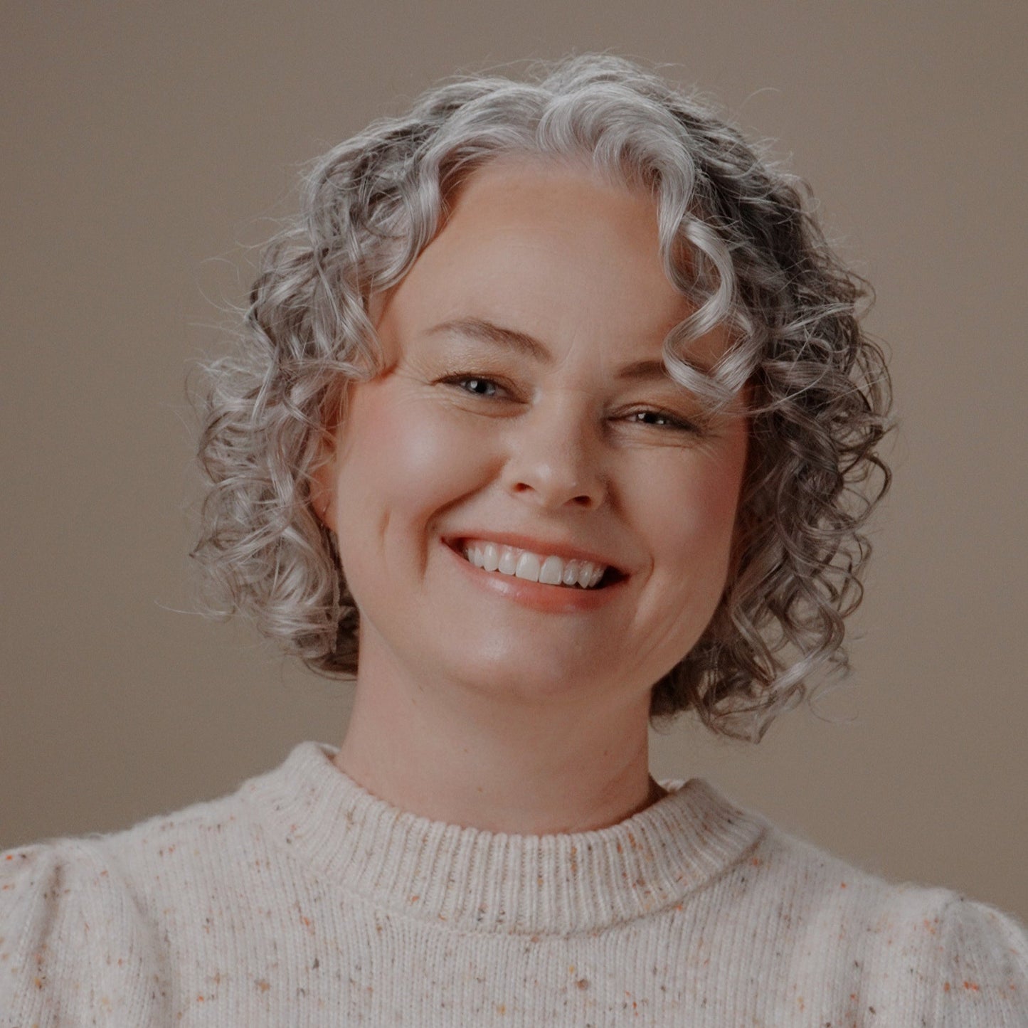 A smiling woman with short curly gray hair wearing a cream speckled knit sweater, photographed against a plain warm beige background.