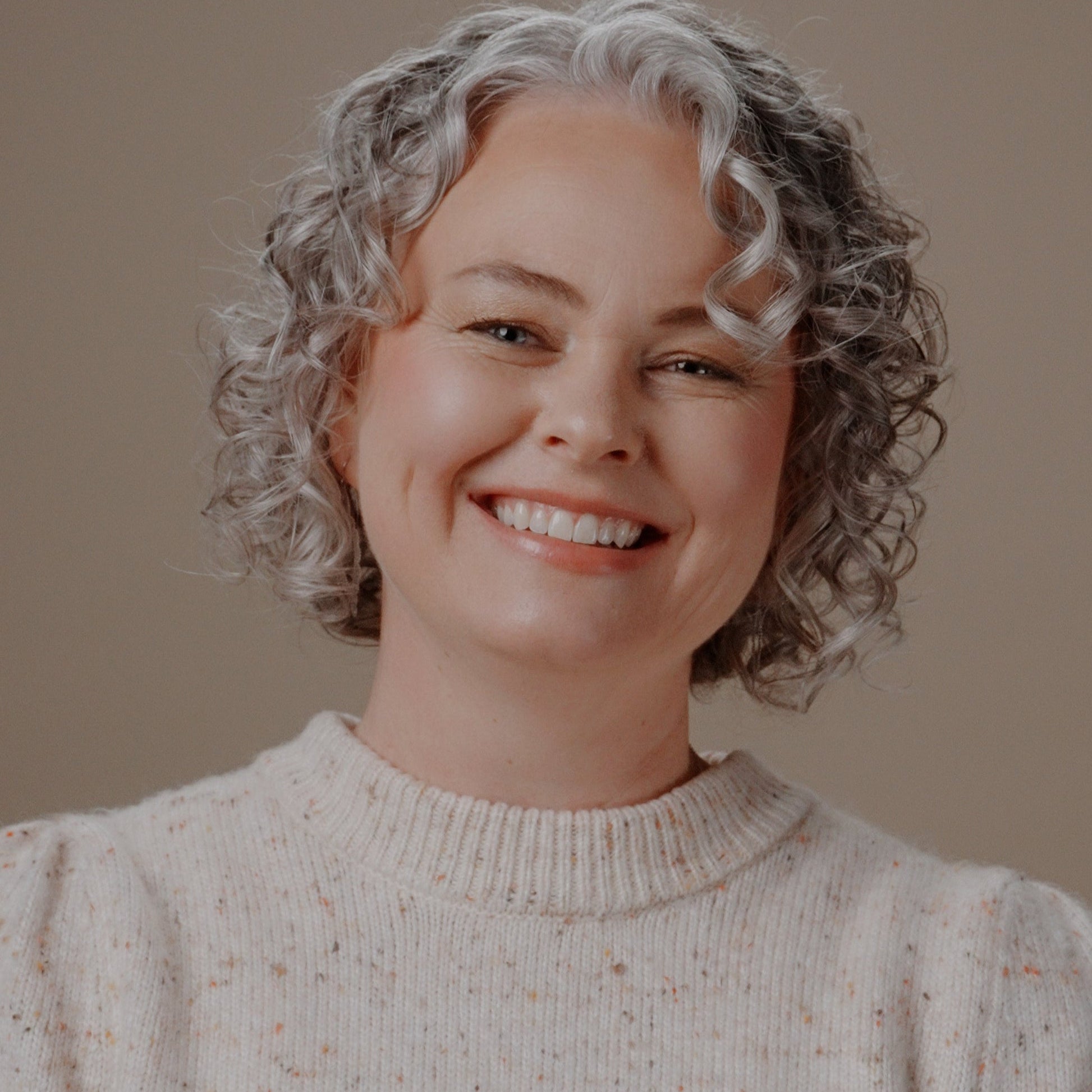 A smiling woman with short curly gray hair wearing a cream speckled knit sweater, photographed against a plain warm beige background.