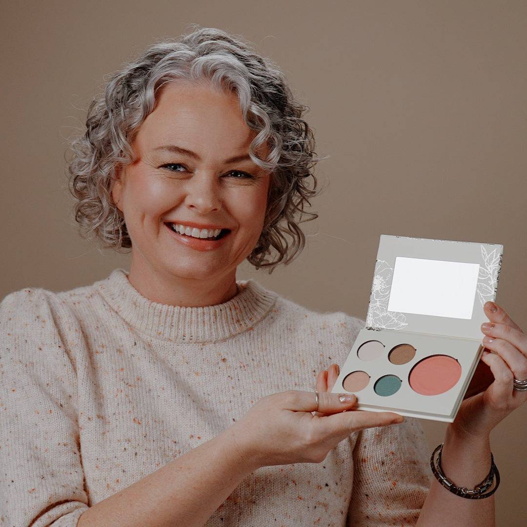 A smiling woman with short curly gray hair holds an open makeup palette with multiple round pans, including a large coral-pink shade, against a plain warm beige background.