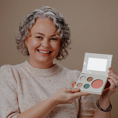 A smiling woman with short curly gray hair holds an open makeup palette with multiple round pans, including a large coral-pink shade, against a plain warm beige background.