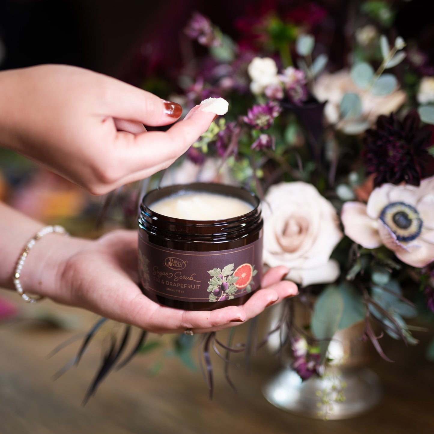 Person holding a jar of cream with a floral arrangement in the background