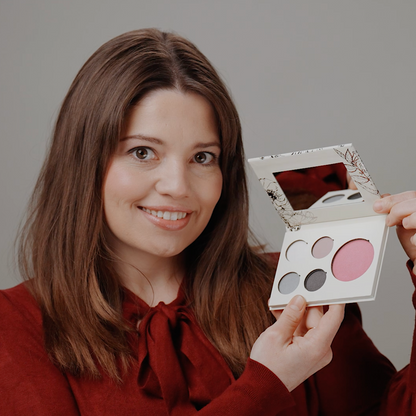 A woman with long brown hair smiles while holding an open makeup palette with four round pans, including pink and gray shades, against a plain gray background.
