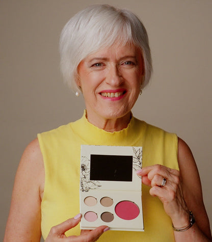 An older woman with short white hair, wearing a yellow sleeveless top, holds up a six-color eyeshadow palette with a mirror. The background is a solid neutral beige studio backdrop.