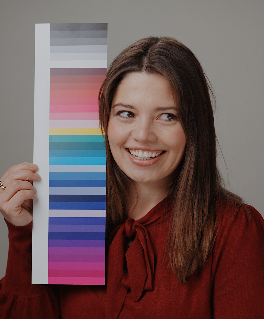 A woman with brown hair wearing a rust-red bow-tie blouse holds up a vertical color swatch card showing a gradient of colors from gray and pink to blue, green, and purple, against a solid gray background.
