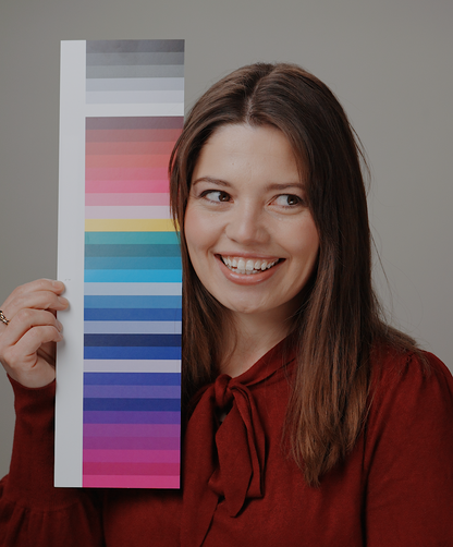 A woman with brown hair wearing a rust-red bow-tie blouse holds up a vertical color swatch card showing a gradient of colors from gray and pink to blue, green, and purple, against a solid gray background.