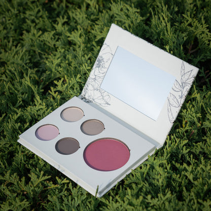 An open makeup palette with four circular and one oval powder pans in neutral and pink shades, resting on green foliage. The compact has a white cover with delicate floral line art and a mirror in the lid.